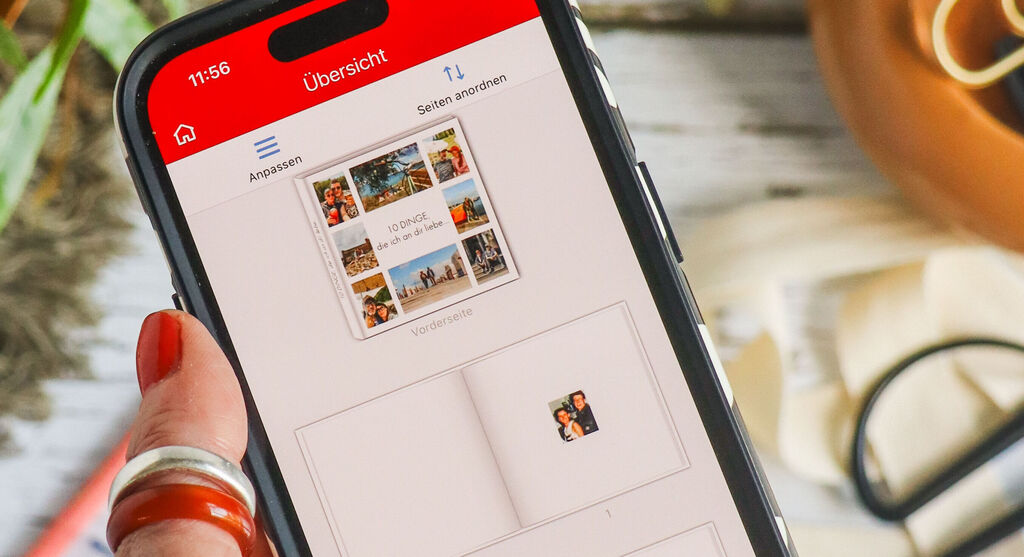 În prim-plan, o mână cu unghii roșii ține telefonul pe care prinde viață, direct în aplicația mobilă CEWE Fotowelt, fotocartea creată anterior. În fundal, pe masă, se vede chiar albumul real, înconjurat de mici detalii care dau farmec proiectului: o plantă verde, role de bandă colorată, pixuri, foarfecă și panglică pentru cadouri.